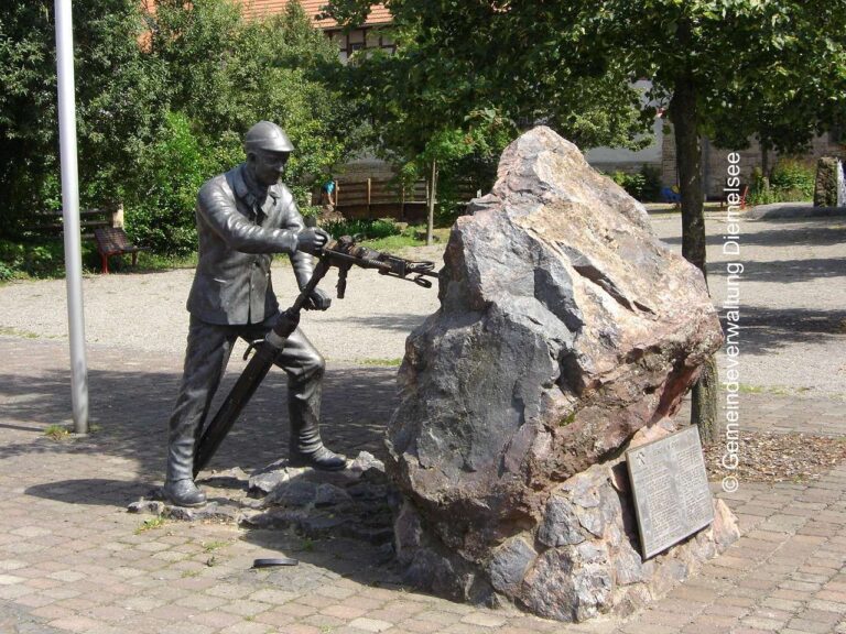Bergamannsdenkmal auf dem Dorfplatz in der Dorfmitte von Diemelsee Adorf. Ein Bergmann aus Guss in Lebensgröße arbeitet mit einem Bohrer an einem großen Diabasstein. Am Stein ist ein Gedenkschild angebracht. Im Hintergrund der Dorfplatz mit Bäumen und Brücke.