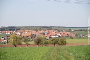 Ortsteil Vasbeck. Einige Häuser mit roten Dächern, in der Mitte die Vasbecker Kirche. Umgeben von Feldern und Wäldern.