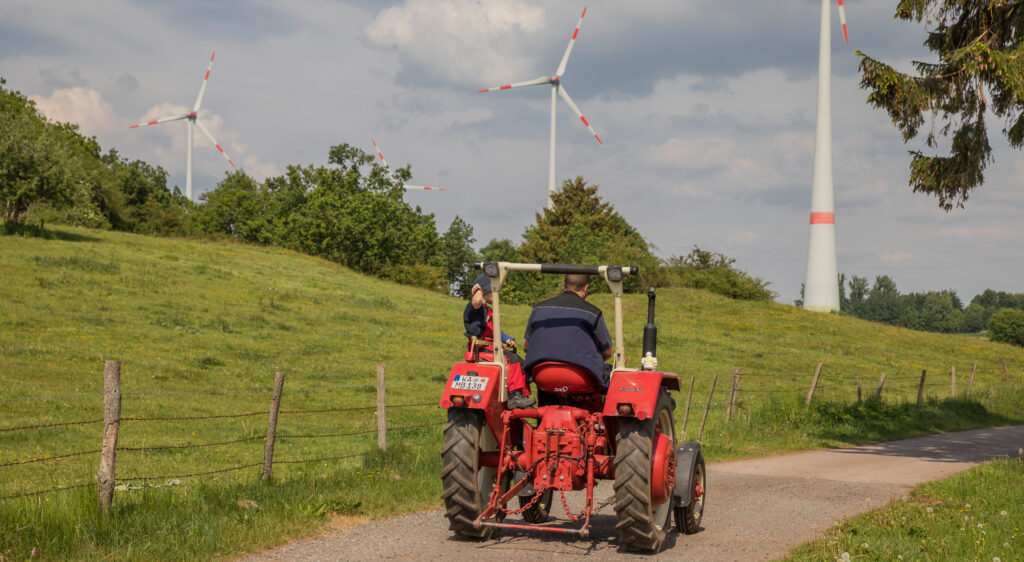 Ein alter, roter Traktor von hinten auf der Fahrt auf einem asphaltierten Feldweg umgeben von grüner Wiese und einigen Büschen. Im Hintergrund 3 Windräder.