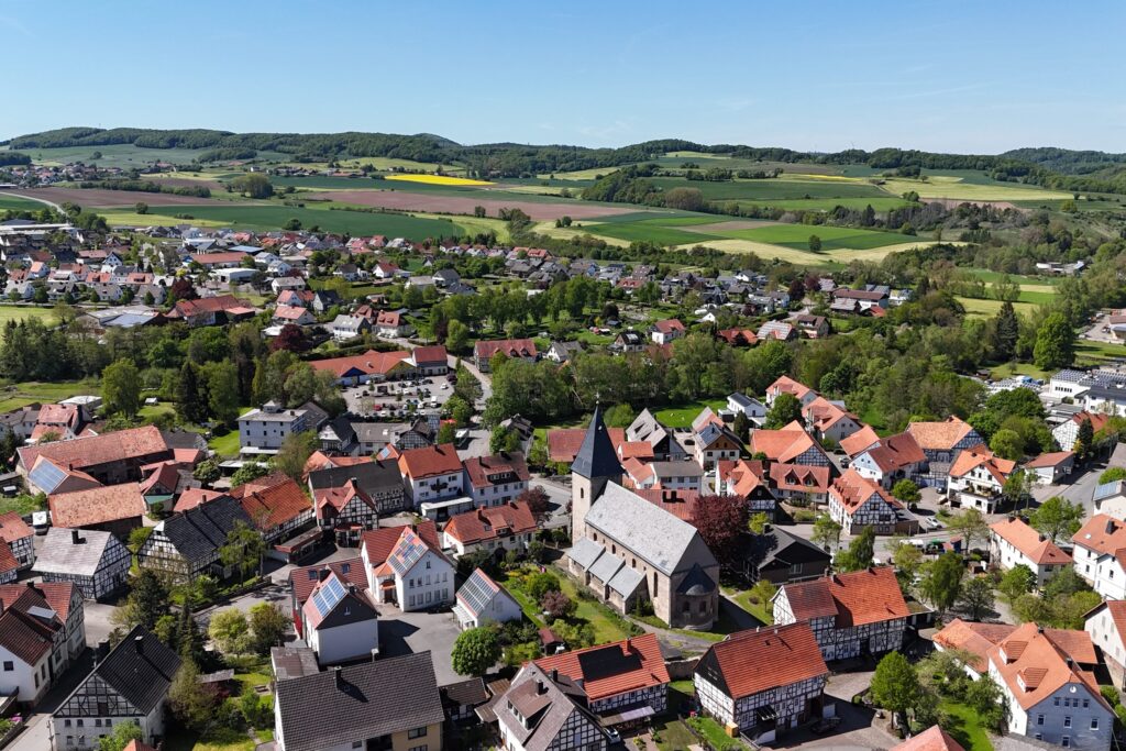 Ortsteil Adorf, Sitz der Gemeindeverwaltung. Ortskern mit der Kirche, rundherum viele Fachwerkhäuser, in der Ferne Neubaugebiet. Blick bis hin zum Nachbarort Rehnegge. Rundherum Wiesen und Wälder.