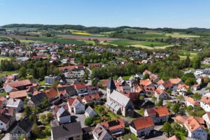 Ortsteil Adorf, Sitz der Gemeindeverwaltung. Ortskern mit der Kirche, rundherum viele Fachwerkhäuser, in der Ferne Neubaugebiet. Blick bis hin zum Nachbarort Rehnegge. Rundherum Wiesen und Wälder.