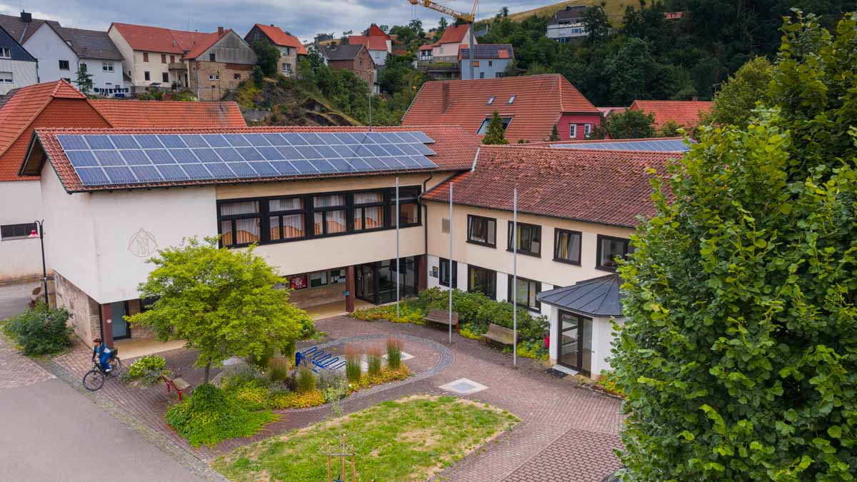 Im Vordergrund ein Beet mit Bäumen und Pflanzen, im Hintergrund ein zweistöckiges Gebäude über Eck gebaut in hellgelb mit braunen Fenstern und rotem Dach.